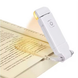 A white handheld book light with a clear clip, shown resting on an open book and illuminating the pages. The device has a power button on the top side and is photographed on a clean white background.