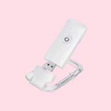 A white USB Book Light with a clear plastic carabiner clip, shown open on a solid light pink background.