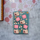 A clear protective case for a Kindle, decorated with a whimsical pattern of pink books, open books, rainbows, clouds, and flowers on a teal background, shown in a flat-lay setting.