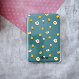 A clear protective case for a Kindle, featuring a printed design of cute cartoon smiling eggs on a teal background, displayed on a textured gray surface with a pink patterned paper corner.