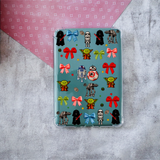 A clear protective case for a Kindle, decorated with a colorful theme print featuring bows on a teal background, displayed on a textured gray surface with a pink patterned paper corner.