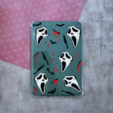 A clear protective e-reader case featuring a Halloween-themed print with white Ghost masks, black knives, bats, and red blood splatters on a muted teal background, displayed on a split pink and gray surface.