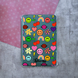 A clear protective tablet case with a colorful retro pattern of smiley faces, cherries, lips, flowers, rainbows, and lightning bolts, shown on a textured gray surface with a pink patterned paper background.