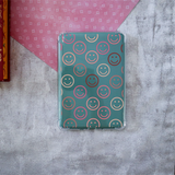 A clear protective case for a Kindle, featuring a printed pattern of colorful smiley faces on a teal background, displayed on a textured gray surface with a pink patterned paper accent.