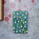 A clear protective case for a kindle, decorated with a colorful pattern of potted succulents, displayed on a gray textured surface next to floral patterned paper.