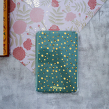 A clear protective case for a Kindle, decorated with a green background and a pattern of yellow flowers and black-and-yellow bumblebees. The case is displayed on a textured gray surface next to a floral patterned sheet of paper.