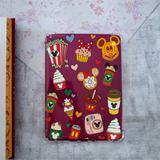 A hard-shell kindle case with a dark magenta background and a collage of illustrations, including popcorn, cupcakes, ice cream cones, coffee cups, and hearts, shown on a gray textured surface with a floral paper fold in the background.