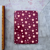 A Kindle case with a glossy dark burgundy background and an allover pattern of pink and white hearts with the words '#1 Mom', shown on a textured gray surface with floral paper in the background.