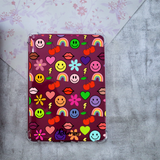 A clear protective case for a Kindle, decorated with a colorful retro pattern of smiley faces, rainbows, cherries, flowers, and lips on a dark red background, displayed on a textured gray surface.