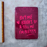 A clear-ridged hardcover Kindle case with a dark magenta-pink back cover, featuring a lined library card-style design with pink handwritten text reading 'BUY ME BOOKS & TELL ME I'M PRETTY'. The case is photographed from above on a gray textured surface with patterned paper in the background.