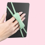 A hand holding a black tablet case with a mint green silicone wrist strap against a solid light pink background.