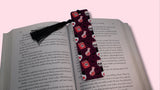 Book with a decorative bookmark featuring skulls, potion bottles, and spell books on a pink background